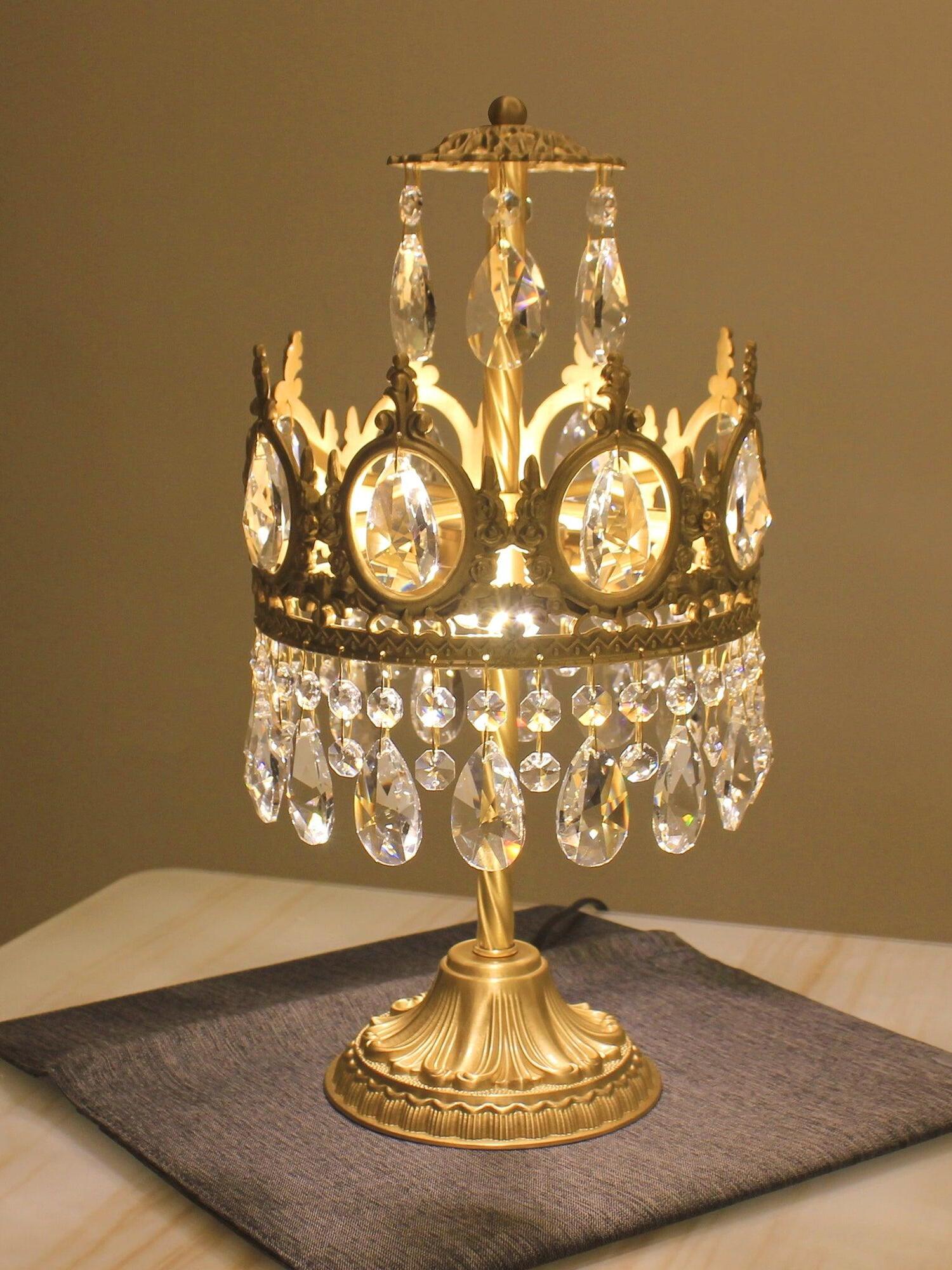 Illuminated crystal table lamp on a dark mat, with an ornate crown and fluted pedestal base.