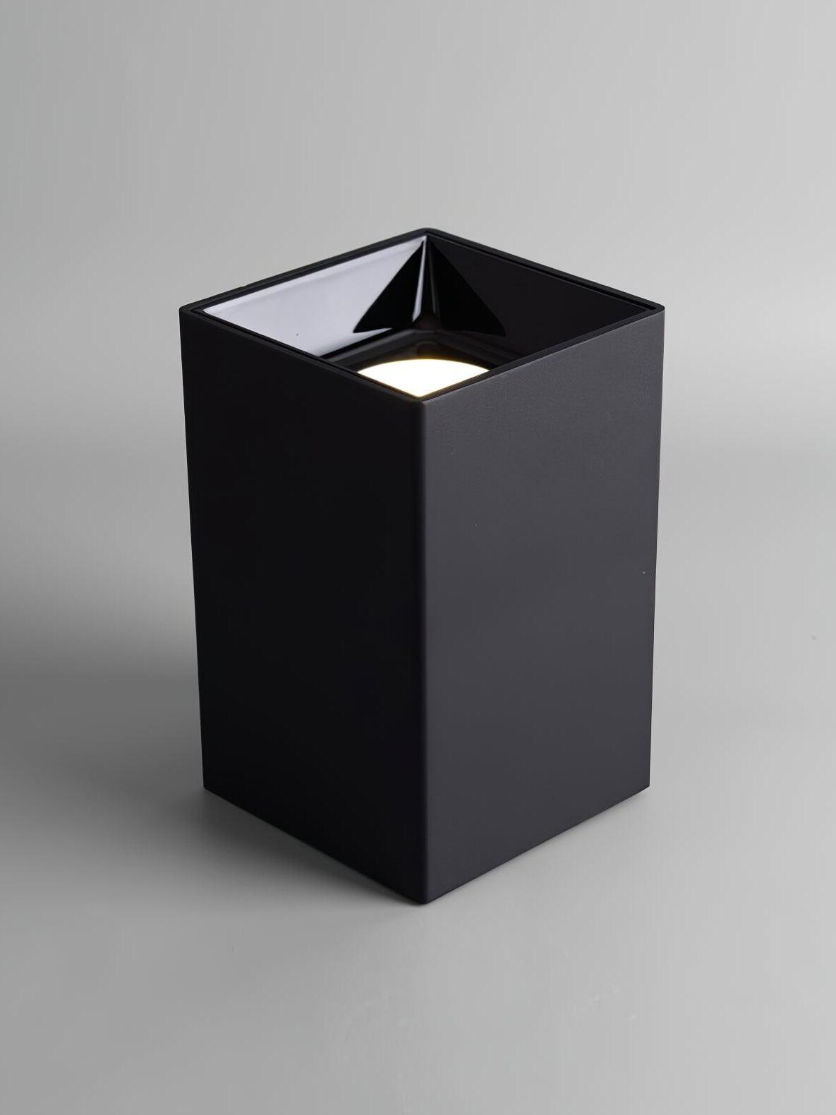Black square spotlight standing with its lit circular aperture facing upward inside a glossy recessed cup
