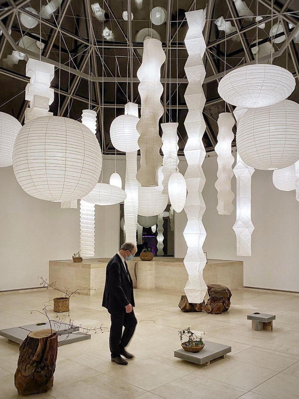 Exhibition hall with many round, flattened, angular and elongated paper lights above displays and a visitor
