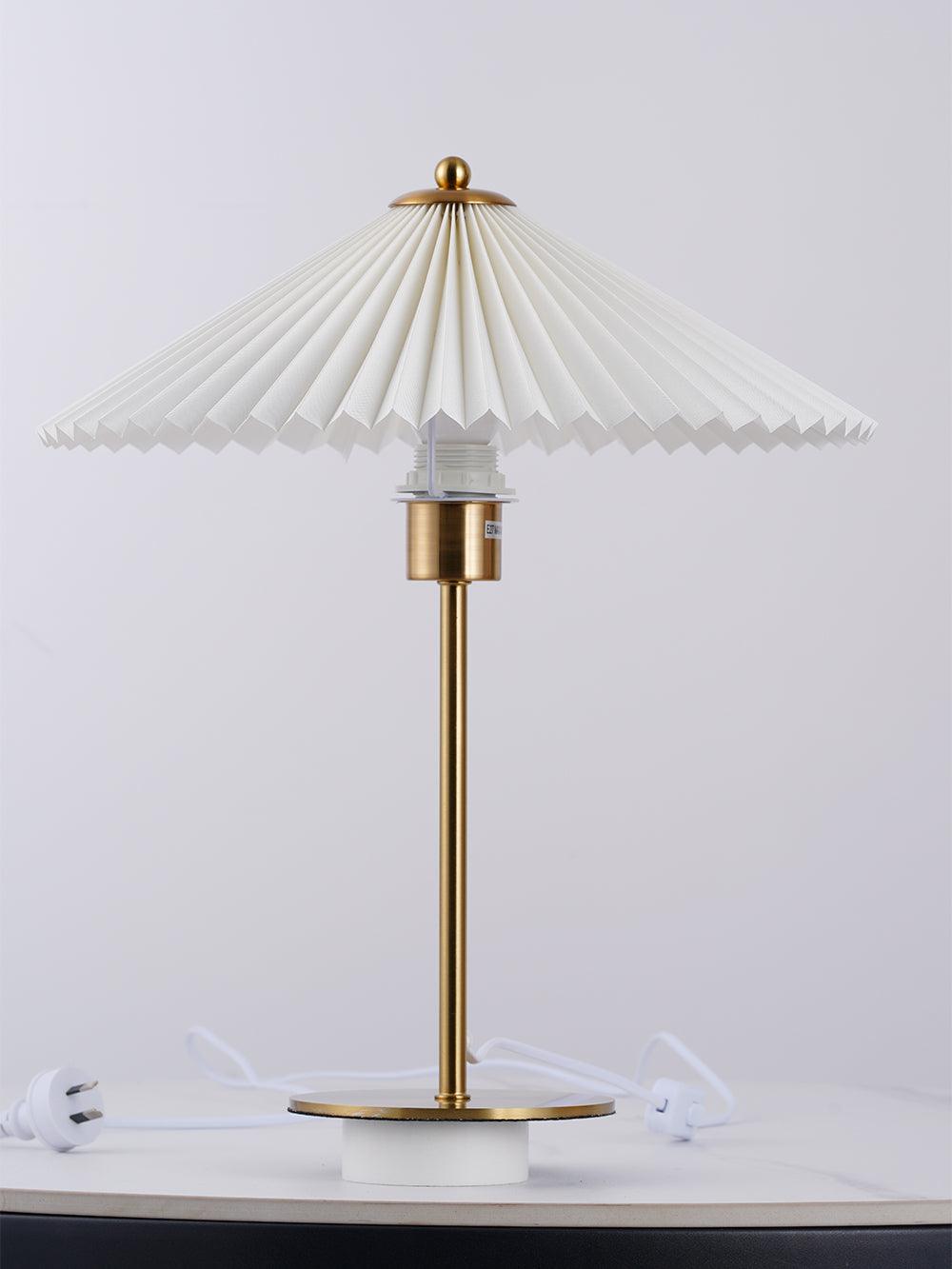 Unlit pleated table lamp on a white surface, showing its gold-tone disk base and white cord