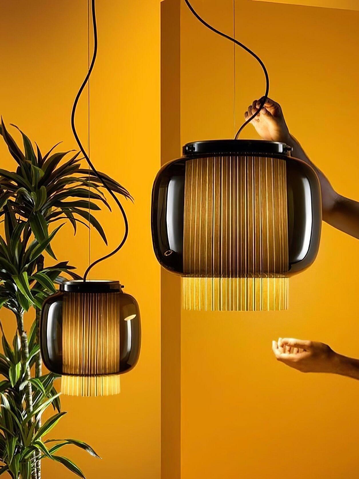 Two smoke-grey Manila T pendant sizes against yellow, with hands beside the larger light and its suspension cable