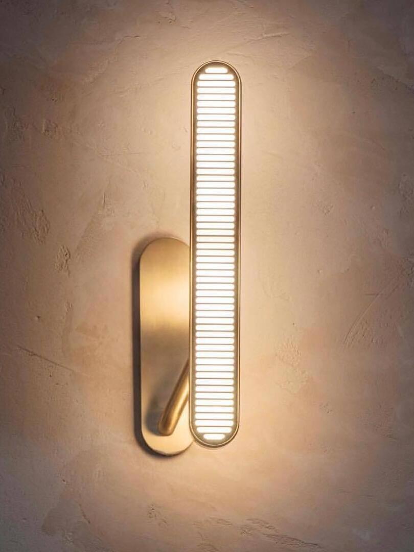 Single lit vertical slotted bar on a gold-tone arm and rounded backplate against a textured Beige wall