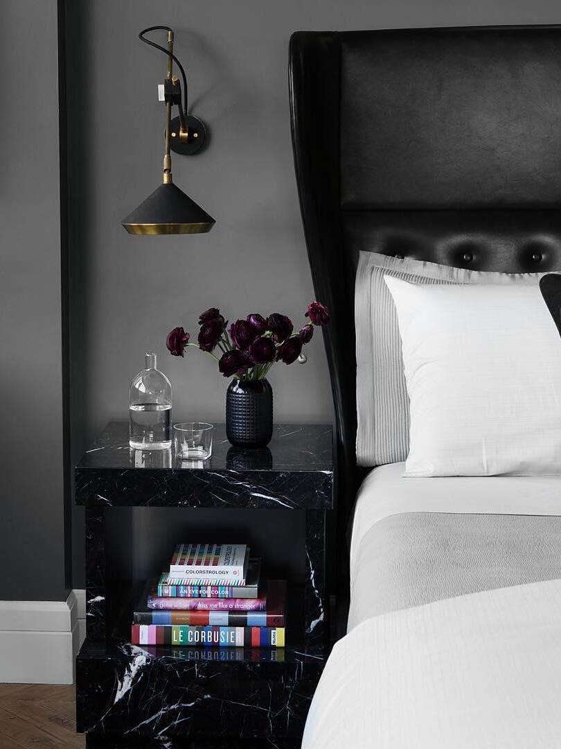 Black cone wall light on a vertical arm above a dark marble-look bedside table and purple flowers