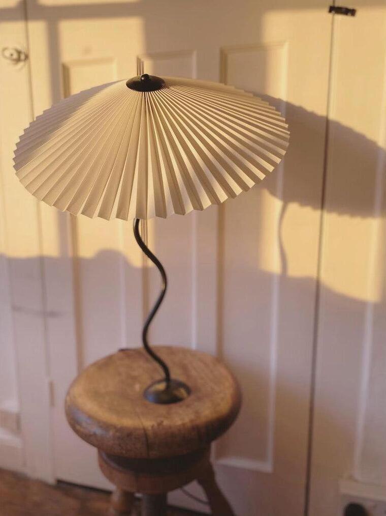 Pale pleated-shade Squiggle lamp on a wooden stool in sunlight, casting a radial shadow on a paneled wall