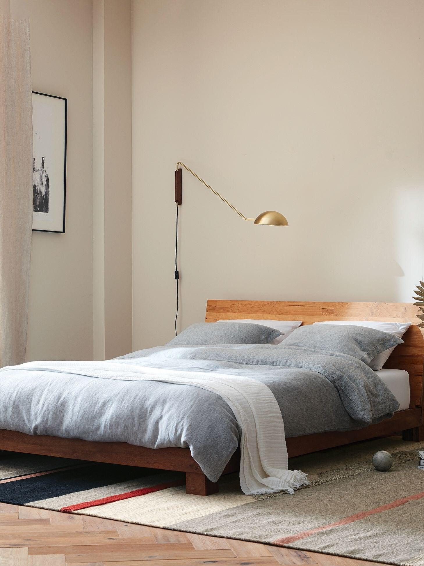 Gold-tone dome wall lamp with long arm and dark wooden mount above a wood bed with grey bedding and switched black cord