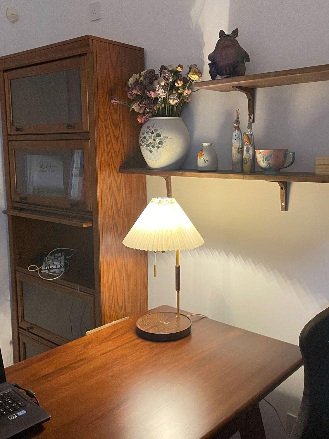 Pleated table lamp glowing on a wooden table beside a cabinet and a wall shelf with flowers