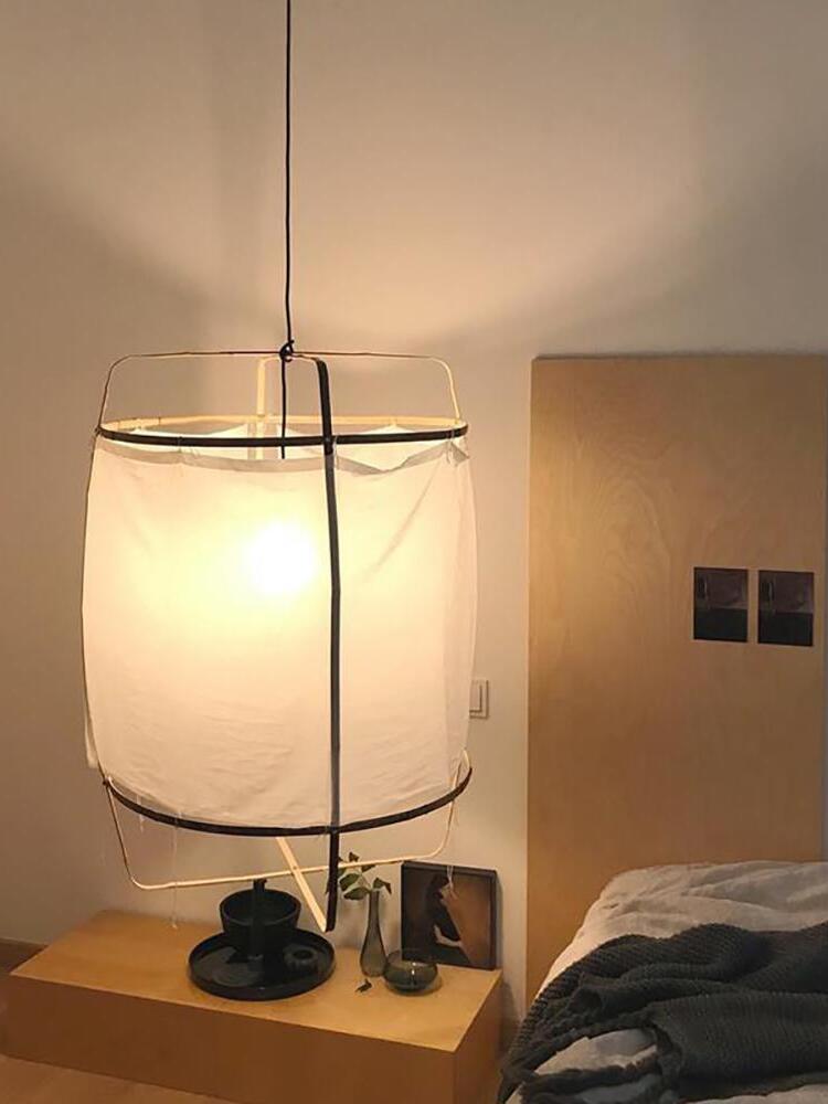 Lit white bamboo lantern pendant beside bed above wooden nightstand with tray, books and a vase