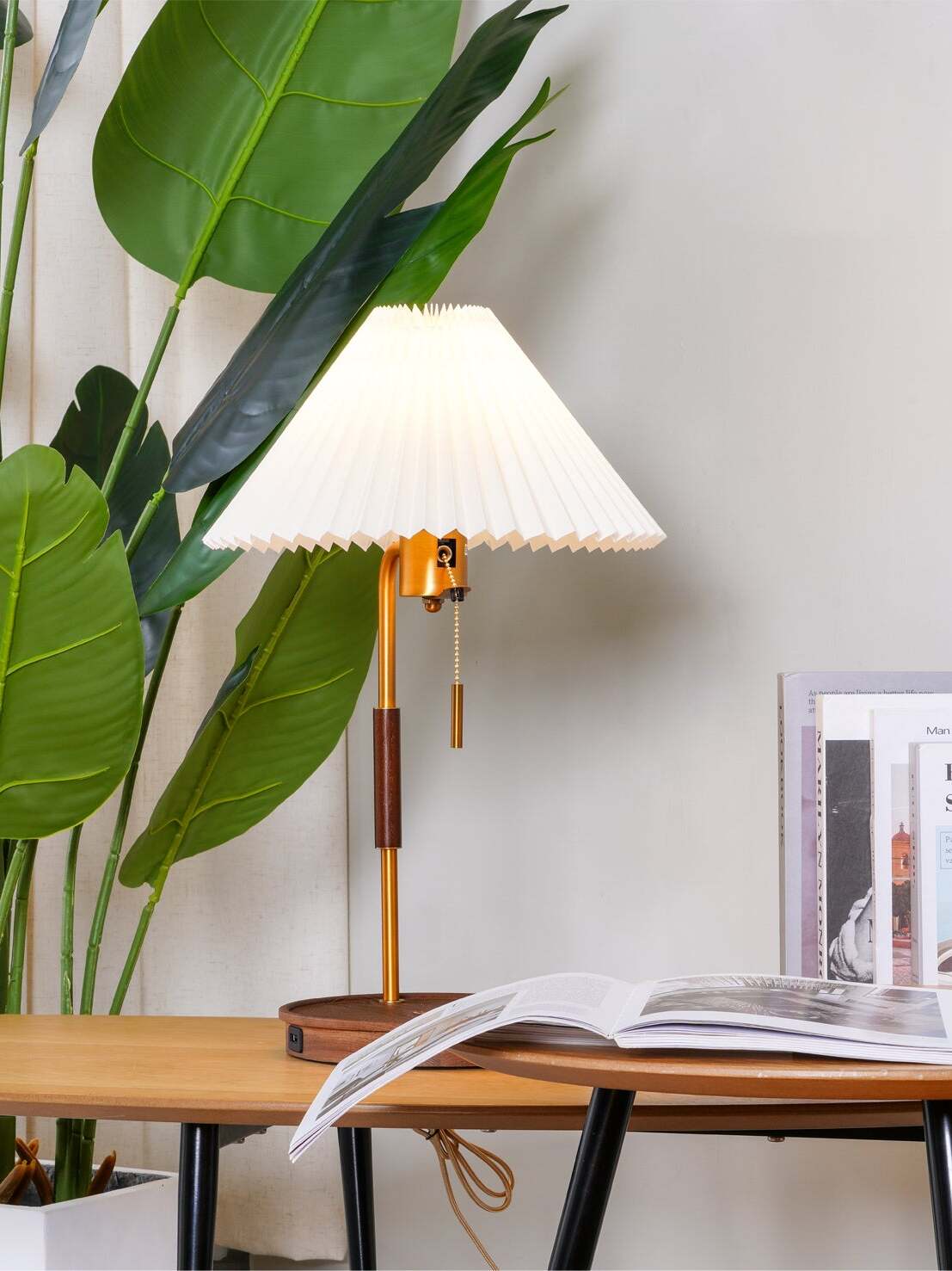 White pleated table lamp glowing beside a leafy plant and an open book on a wooden desk
