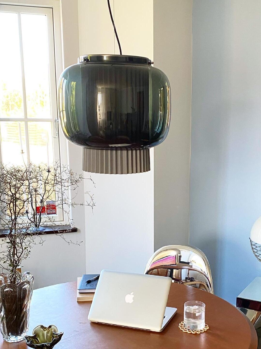 Smoke-grey Manila T pendant light above a round wood worktable with a laptop and branch vase near a window
