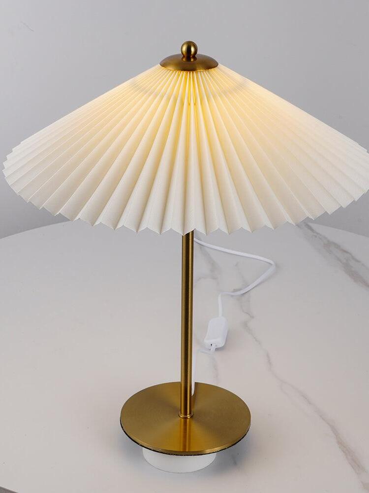 Higher view of the lit pale pleated shade, thin stem and disk base with a white cord behind