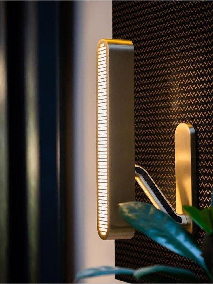 Angled side view of a lit vertical grille bar, gold-tone arm with dark inset and rounded backplate beside Green leaves