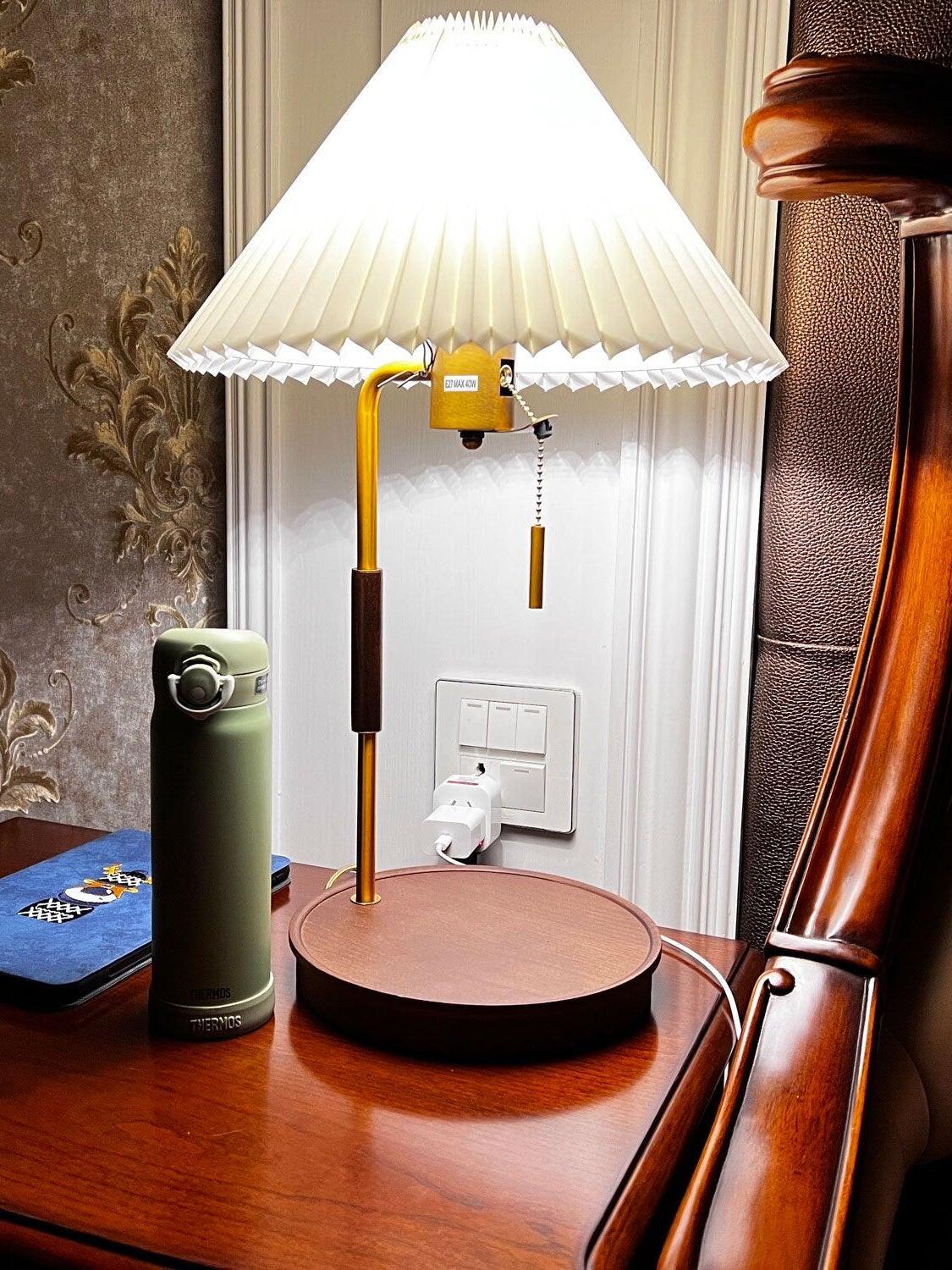 Lit white pleated lamp on a wooden surface beside a green bottle, wall outlet and curved wooden furniture