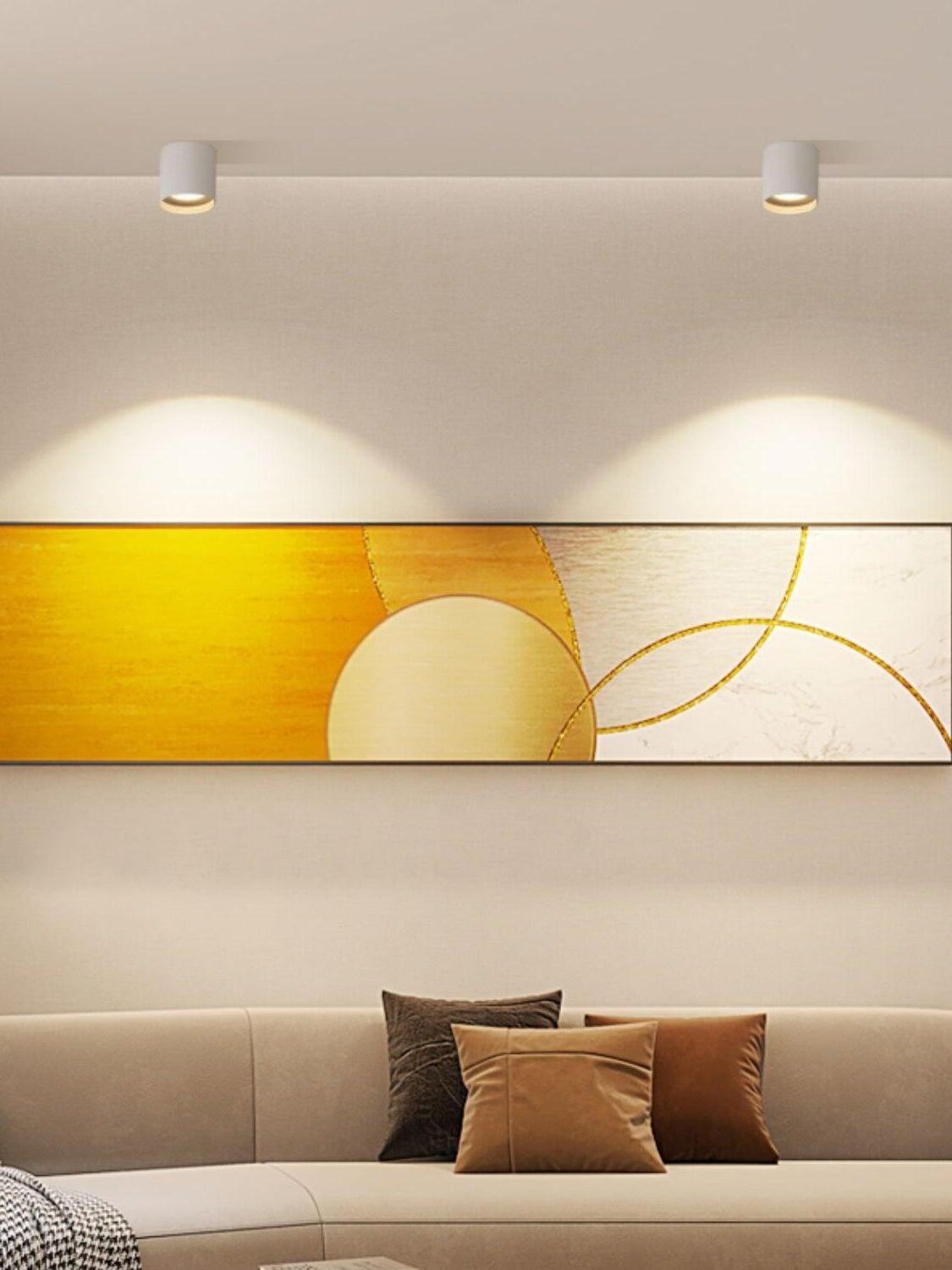 Two white ceiling lights above broad yellow abstract artwork and a pale sofa with cushions