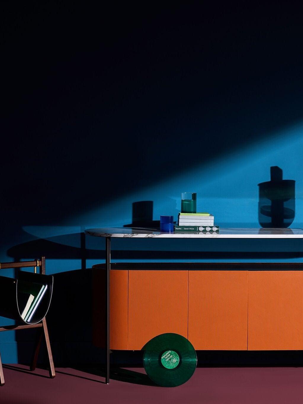Orange cabinet under blue wall lighting with dark decorative vessels, books, a blue cup and a chair