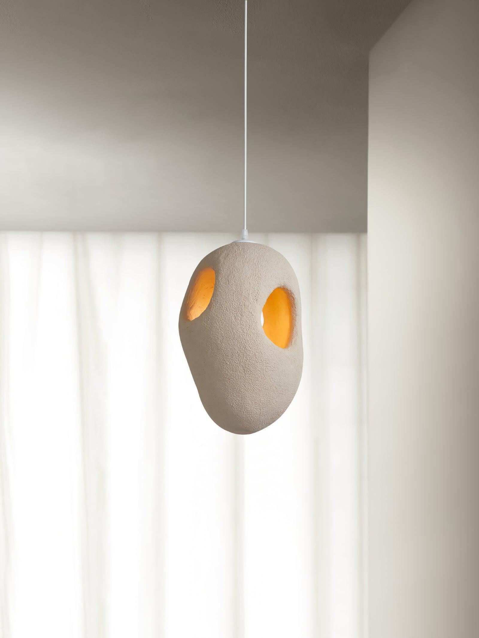 Close-up of a rounder cream-looking textured pendant with two smaller openings glowing warm before sheer White curtains