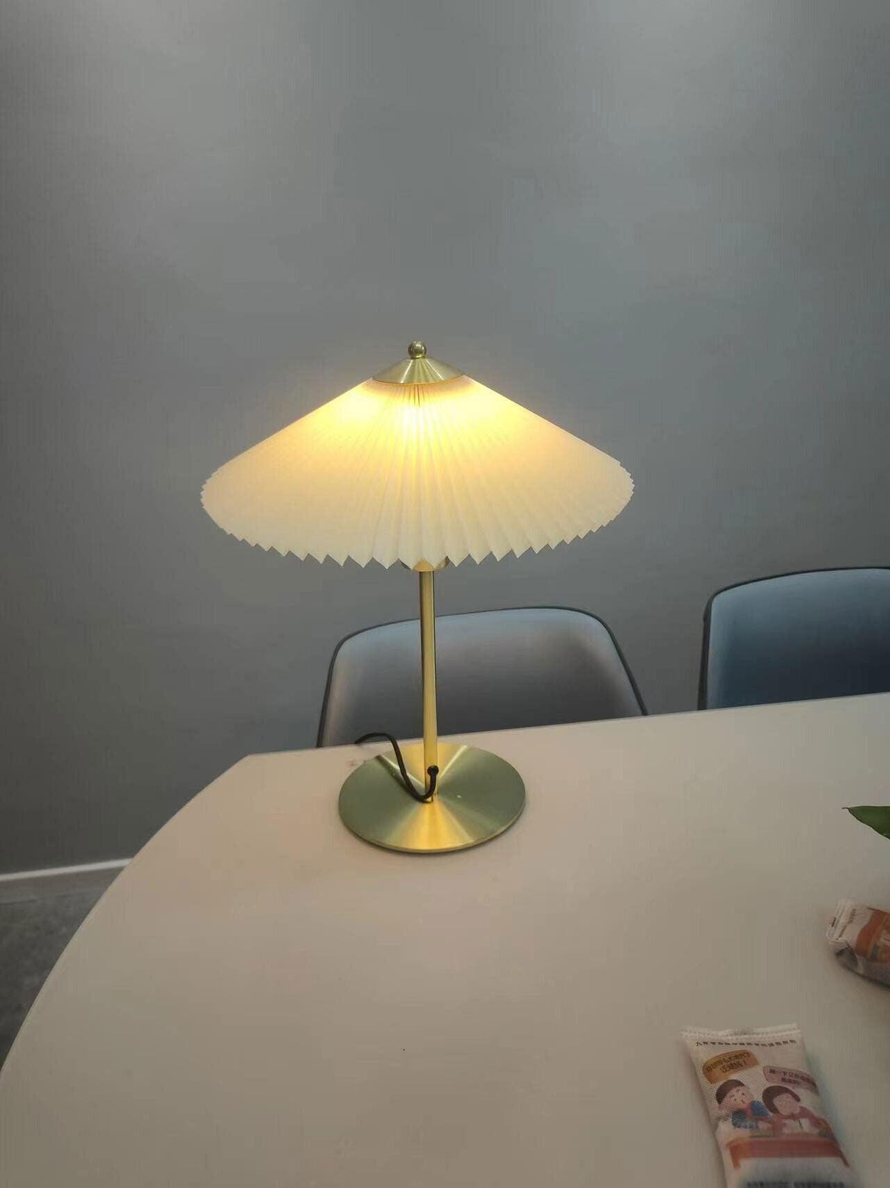 Pleated table lamp glowing on a light-coloured table with two chairs against a grey wall