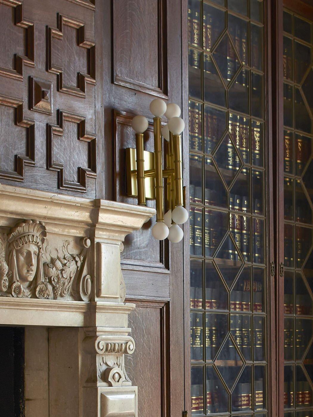 Gold-tone Meurice wall light on a wood panel beside a carved fireplace surround and a glazed bookcase