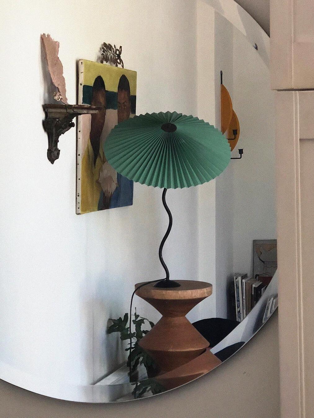 Green pleated-shade lamp reflected in a round mirror, on a wooden stool near artwork and plants