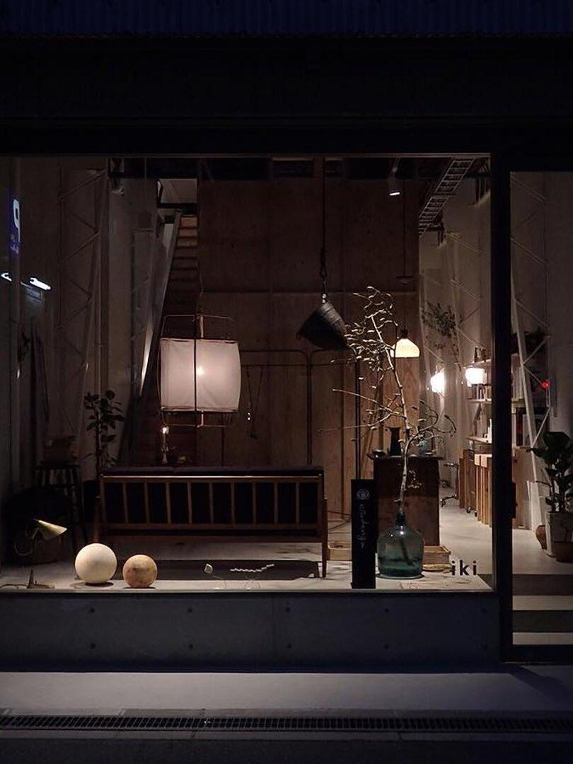 White lantern pendant glowing inside a shop display at night, with wooden ladder, bench, branches and vases