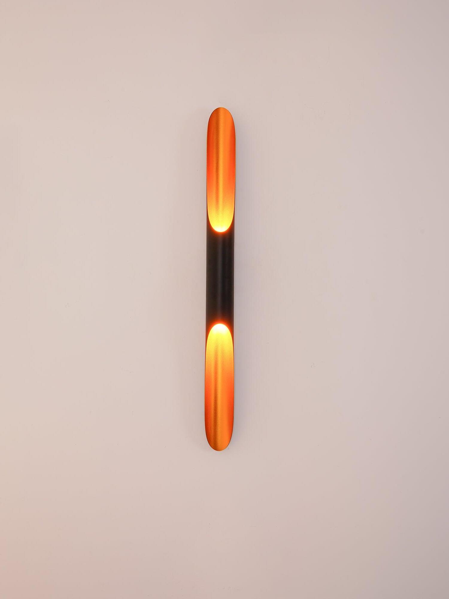 Front view of a single black tube wall light with long glowing gold openings on a white wall