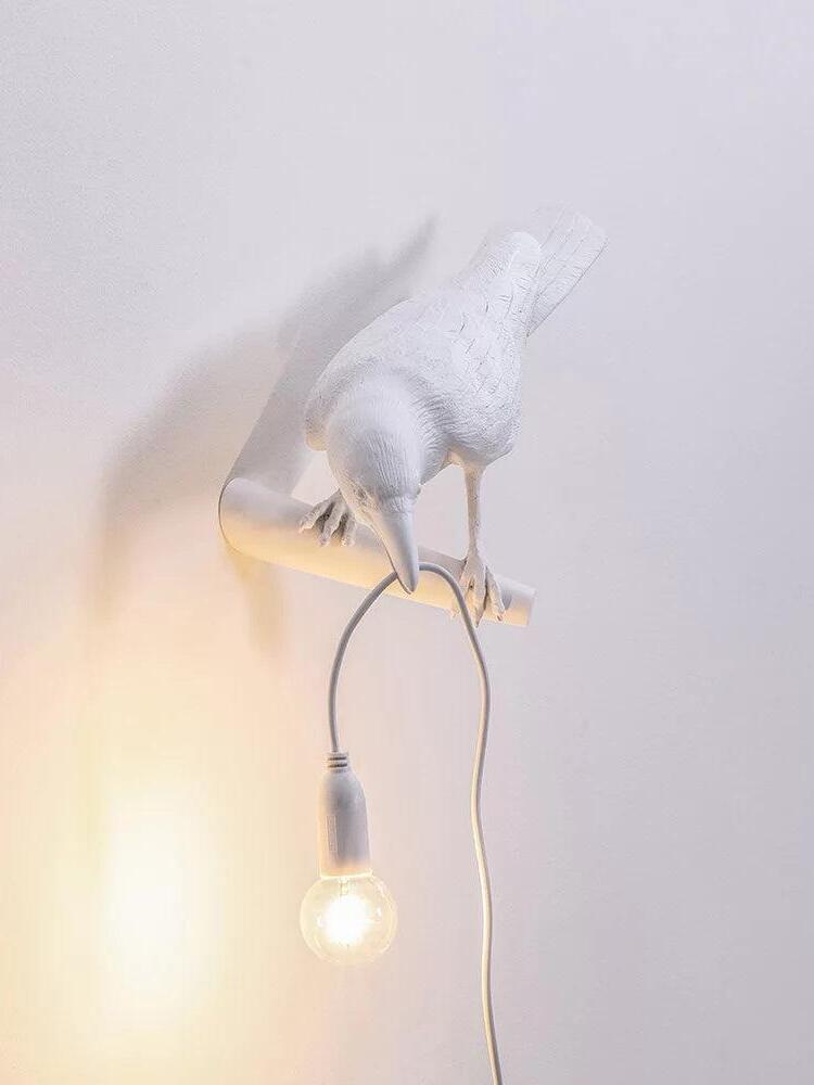 Front-oblique white bird wall light with a hanging lit bulb and cable below the perch