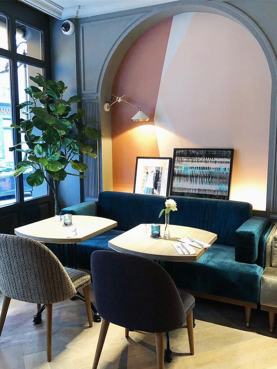 White cone wall light above blue dining seating and tables in a pink arched alcove beside a window