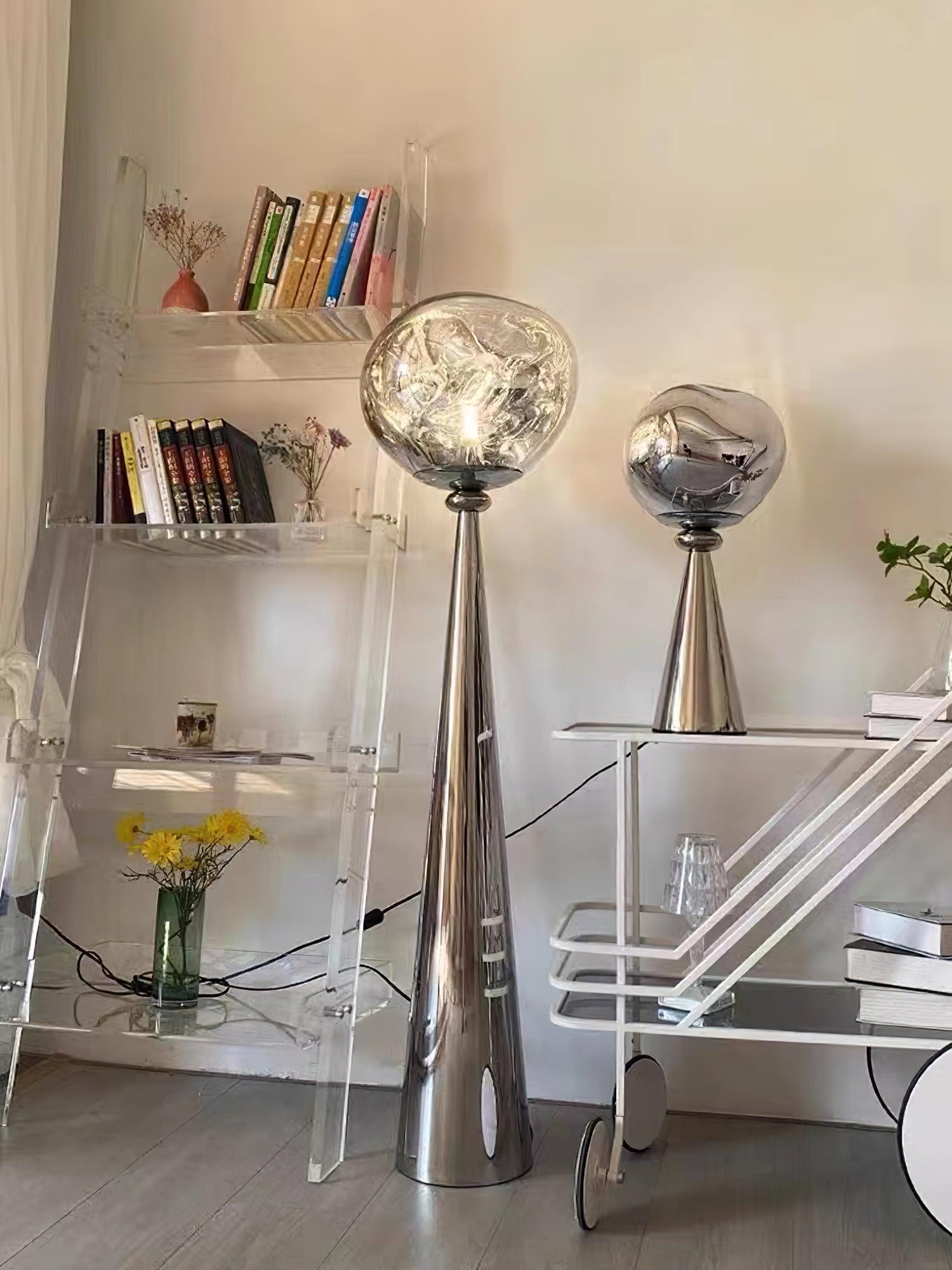 Lit chrome floor lamp beside an unlit mirrored globe table lamp on a white cart by clear bookshelves
