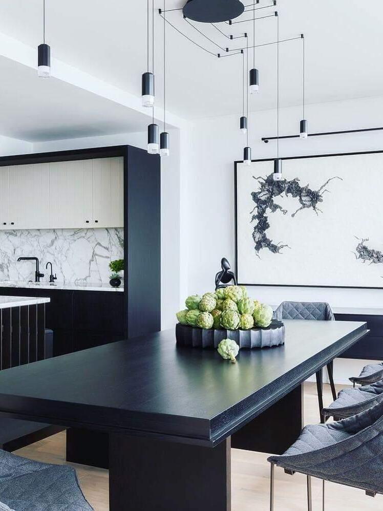 Black cable pendants over a dark dining table beside a kitchen and framed artwork