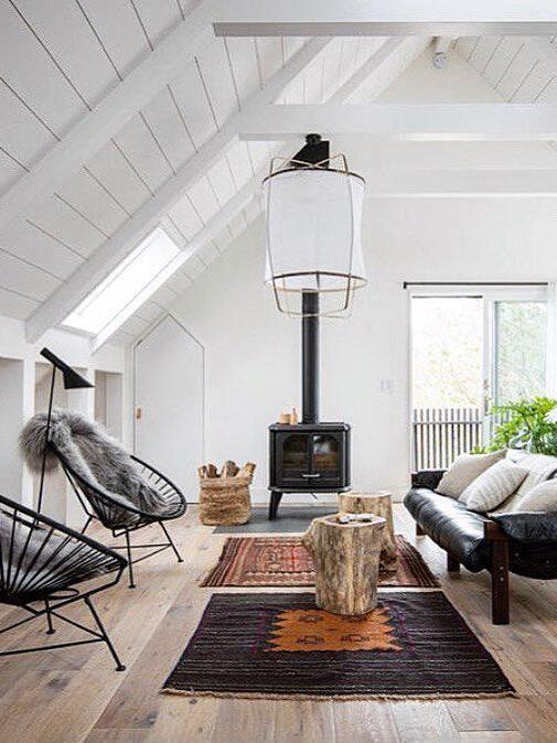 White lantern pendant in a room with pitched beams, black stove and sofa, rustic wooden table and patterned rugs