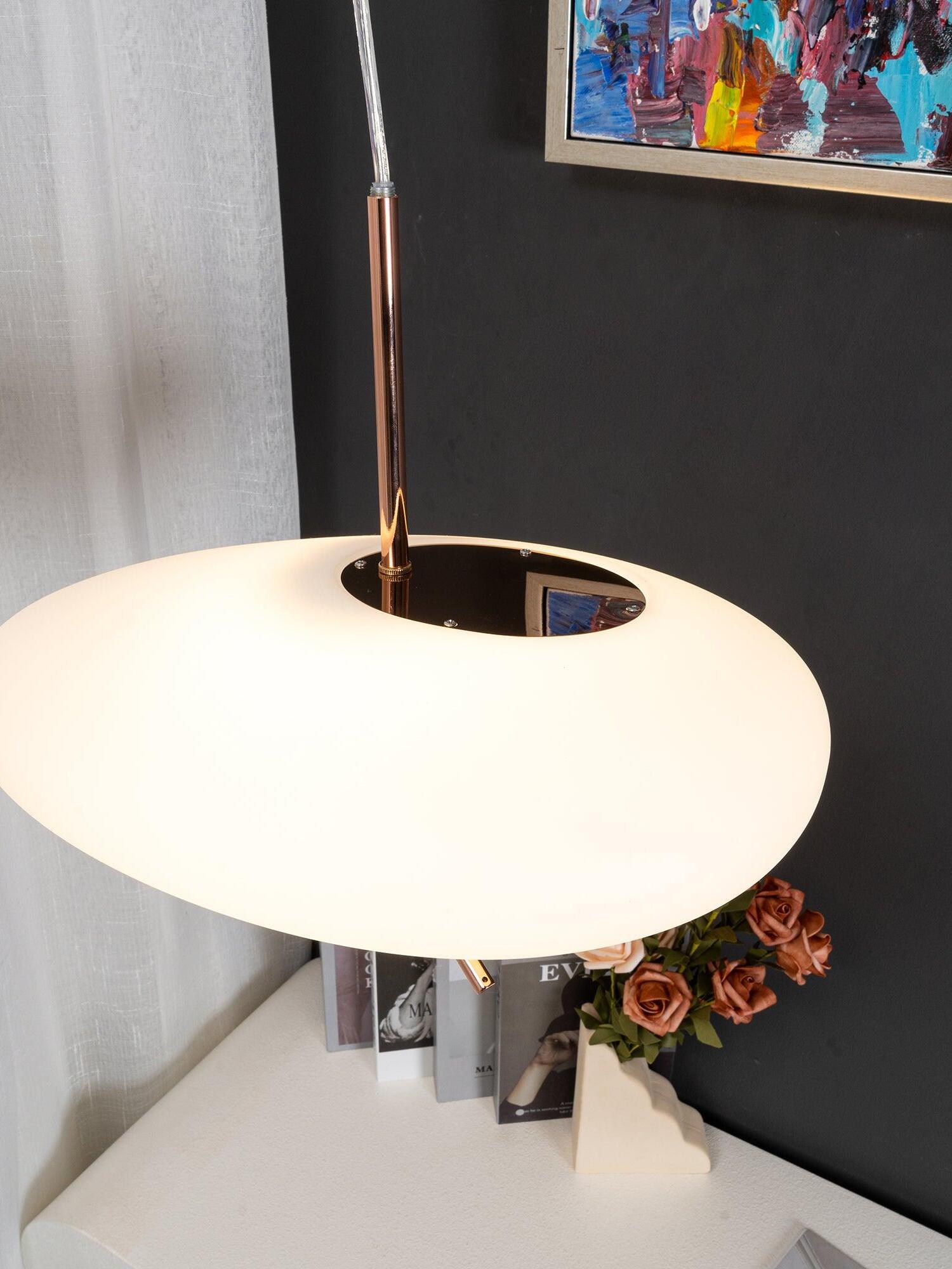 Top close-up of a white shade, reflective central plate and rose-gold stem beside framed artwork