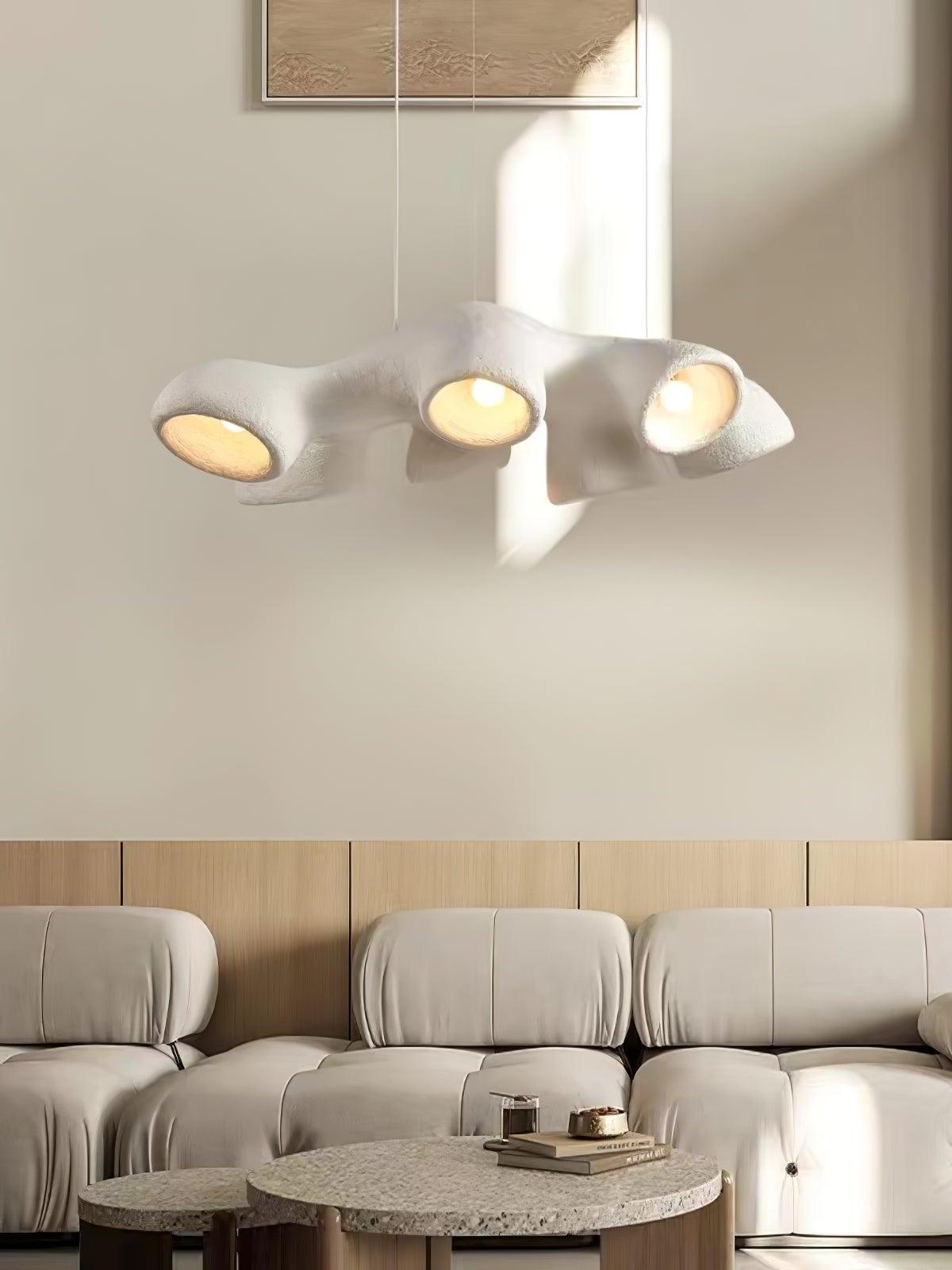 White elongated branching pendant above a pale tufted sofa and round tables beside sunlit wall art