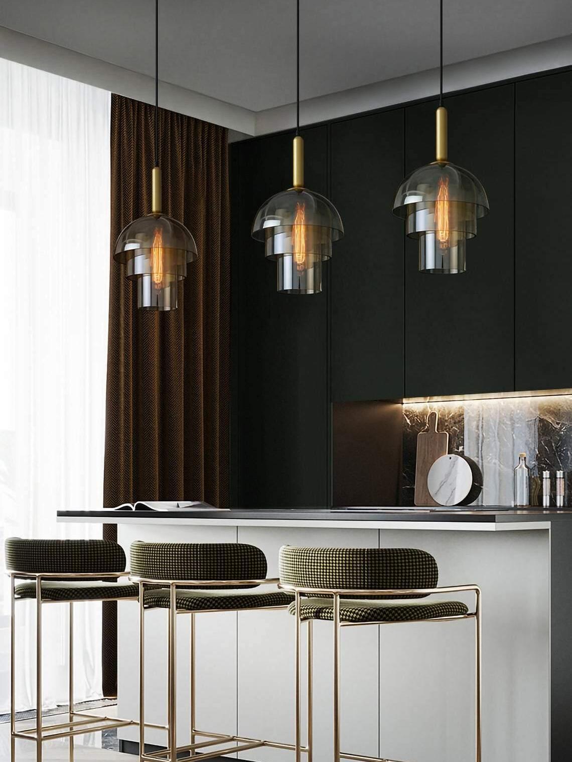 Three tiered glass pendants over a kitchen island and upholstered stools, against dark cabinets and a stone backsplash