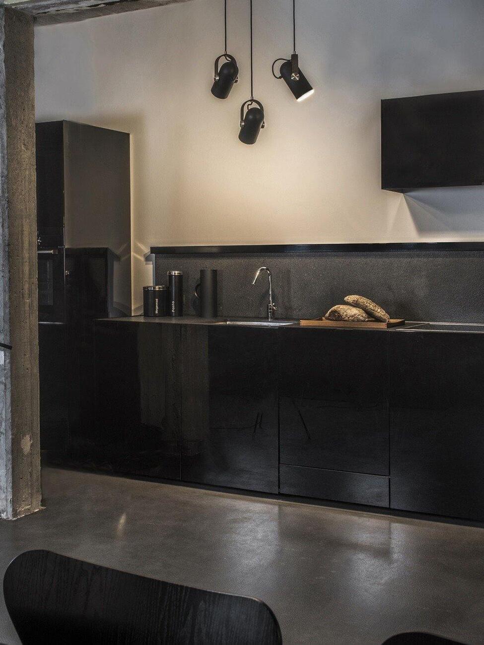Three black pendant heads tilted above a dark kitchen counter and sink, with bread, wall screen and exposed beam
