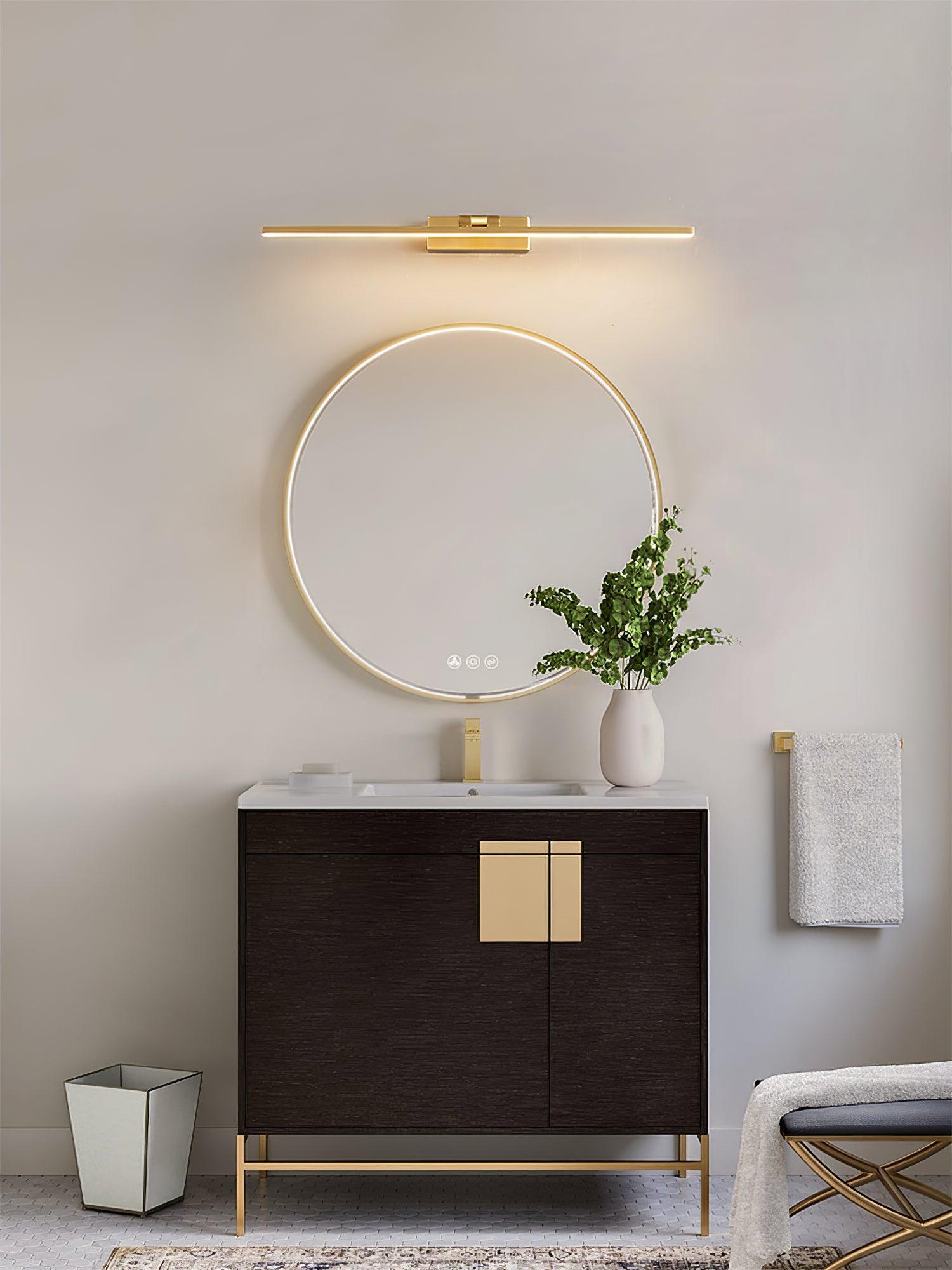 Gold-tone lit wall light above round illuminated mirror, white basin and dark cabinet with vase and towel beside it