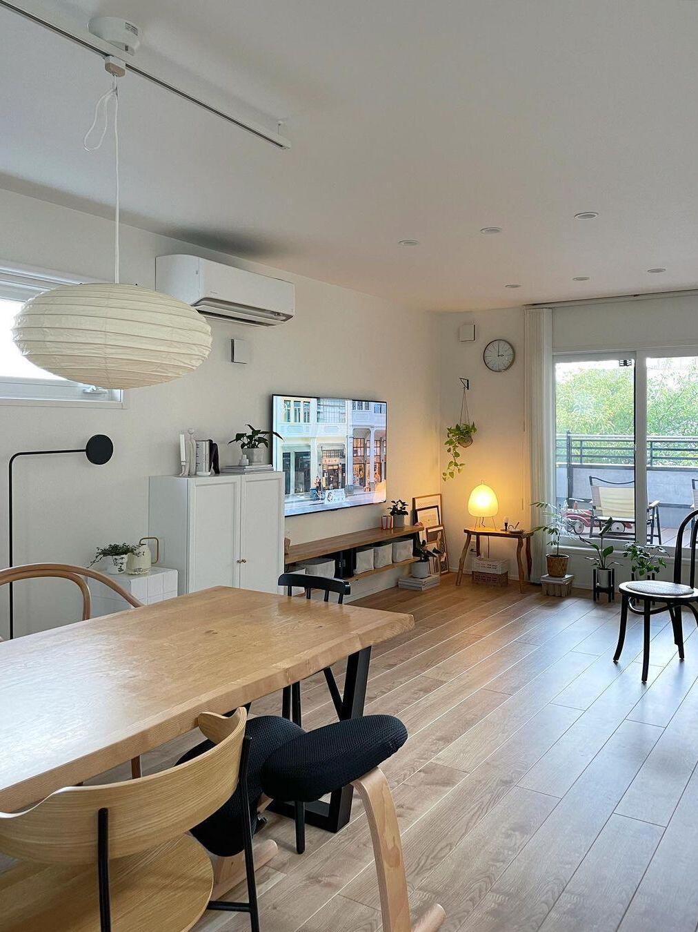Pale paper lantern over a wooden dining table in a white living room, with ceiling track, wall TV, plants and balcony door