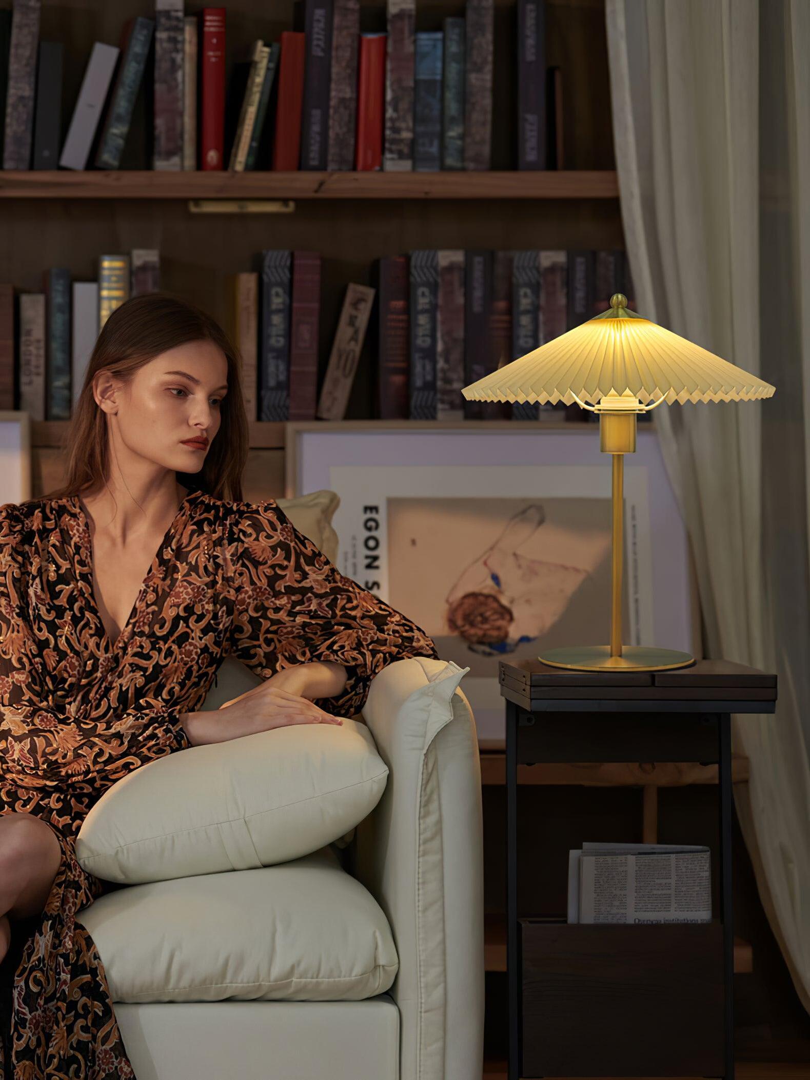 Gold-tone pleated table lamp beside an armchair in a room lined with bookshelves