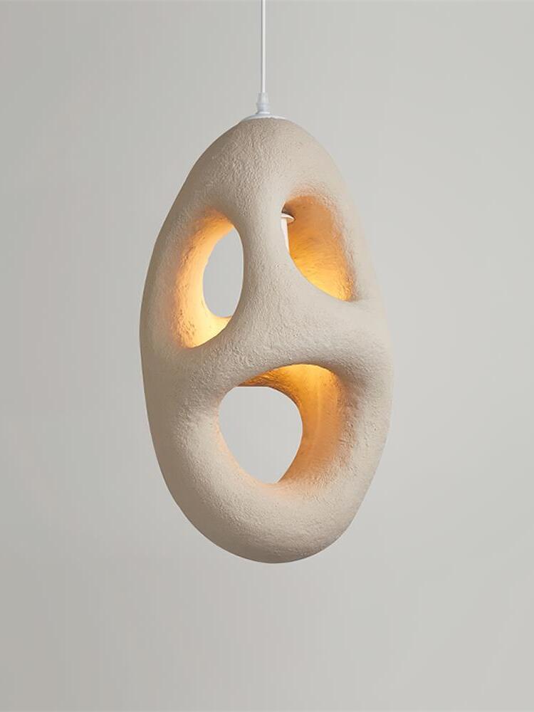Elongated cream-looking textured pendant with three openings glowing warm inside on a White cord against Gray