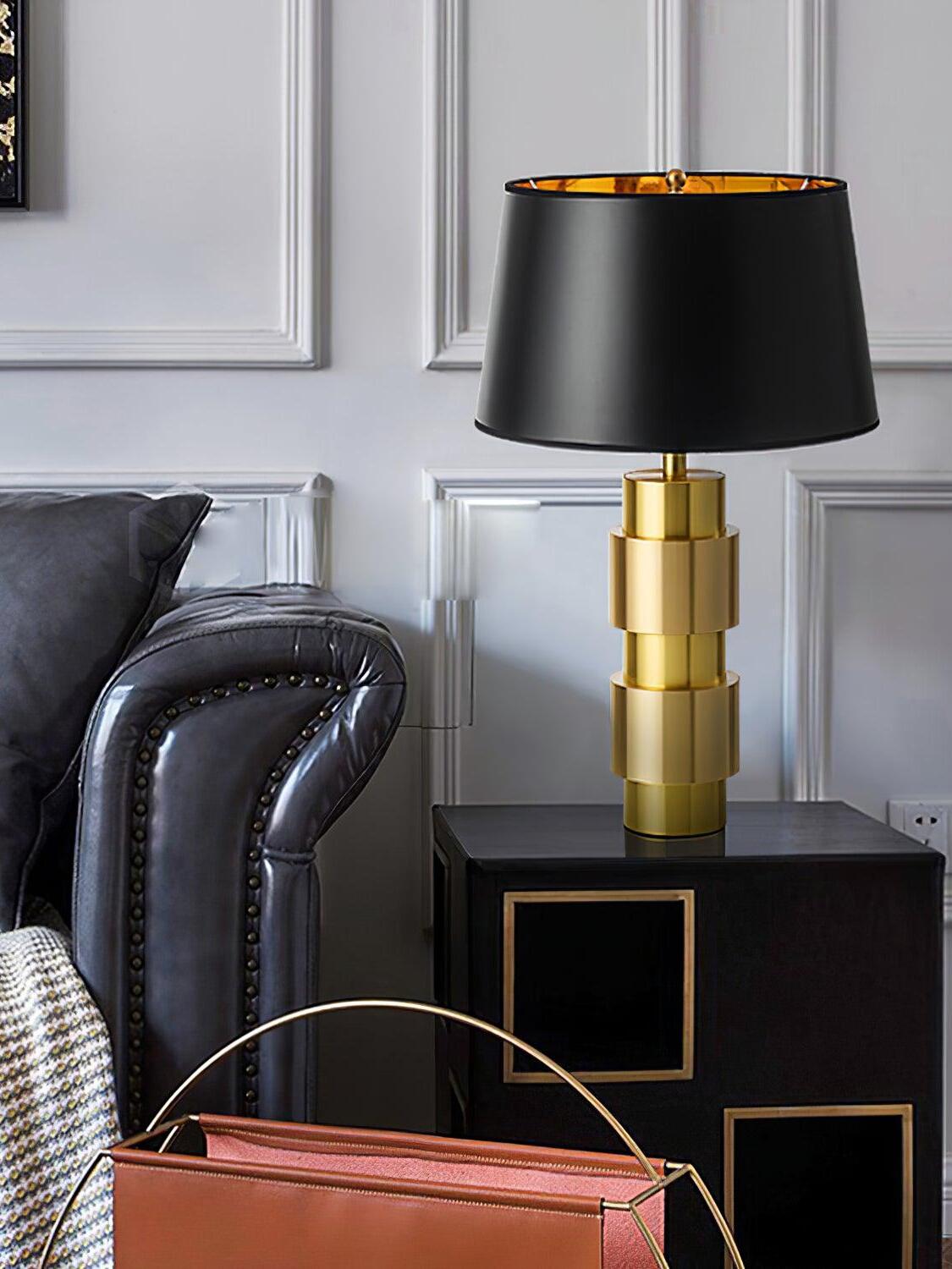 Gold-tone cylindrical lamp with a black shade on a dark side cabinet beside a leather chair