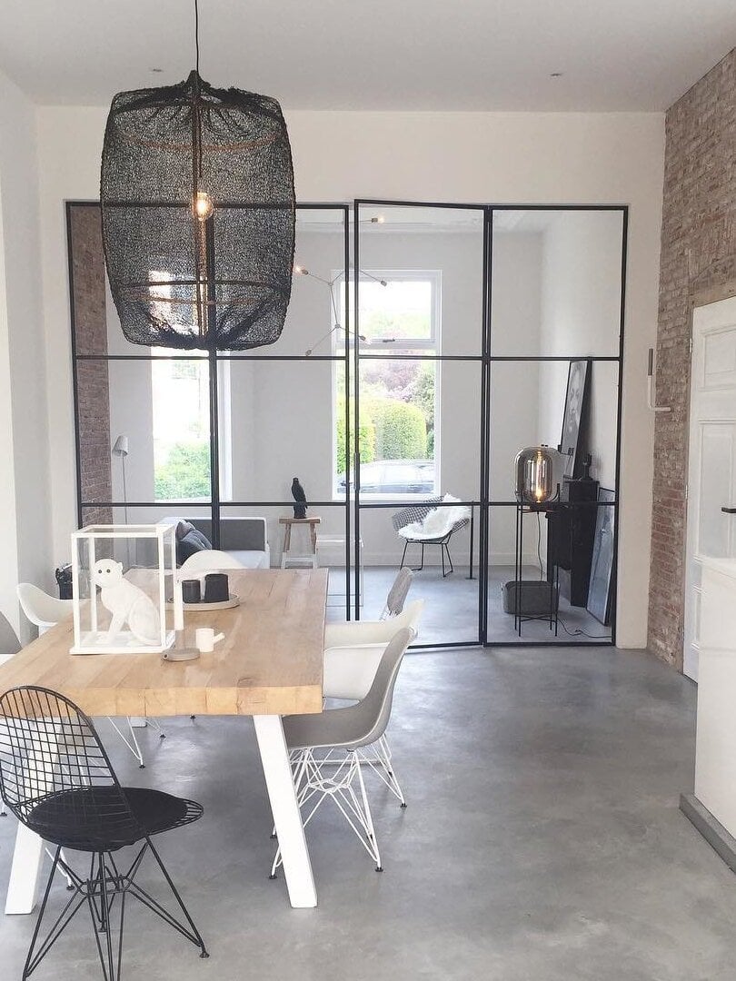 Black open-mesh lantern shade over bamboo ribs above dining table, with visible bulb, glazed partition and brick wall