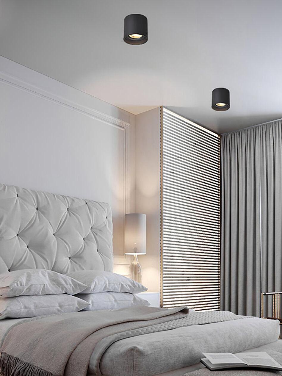 Two black cylindrical ceiling lights above an upholstered bed, with bedside table lamp, blinds and grey curtains