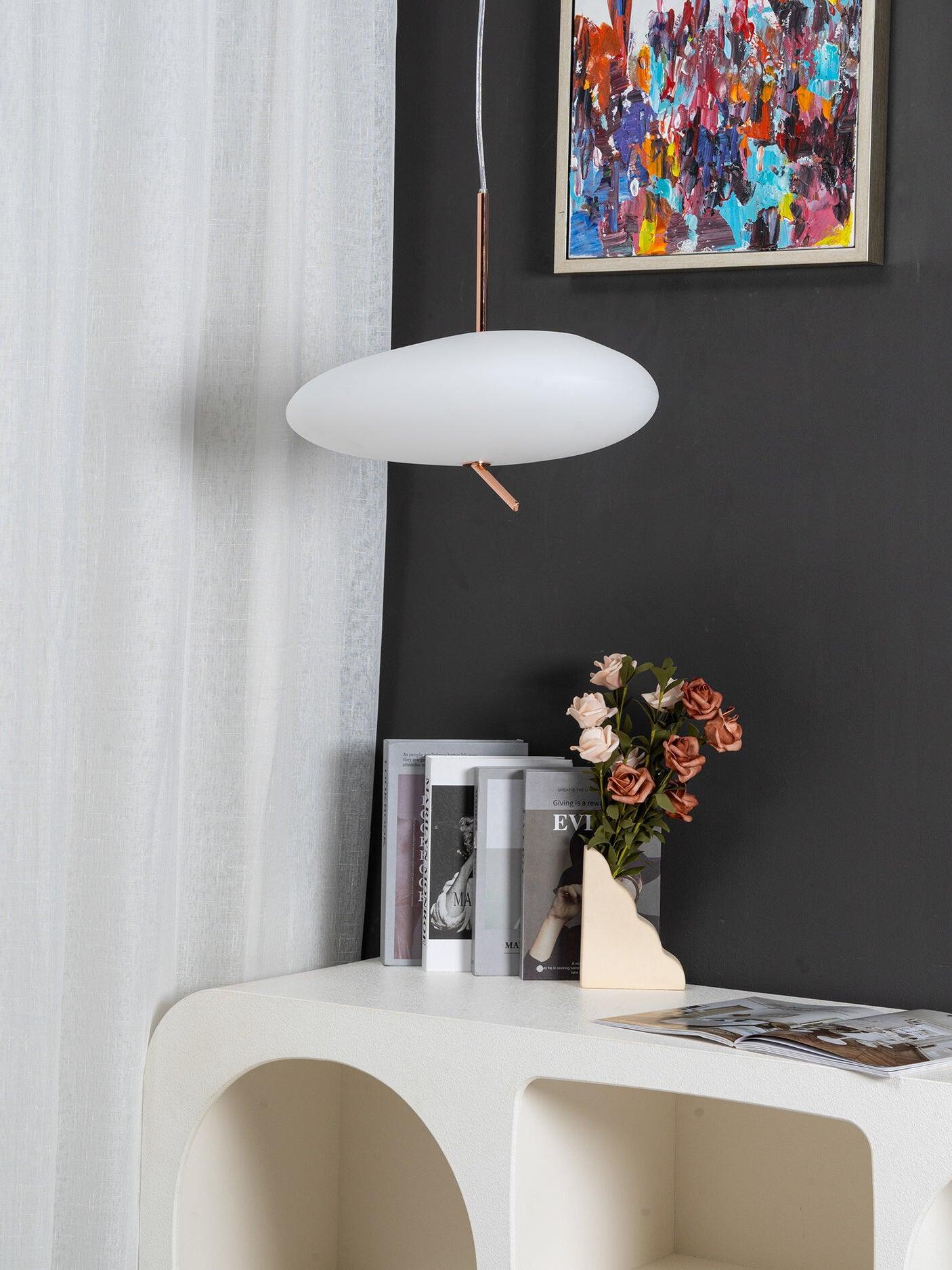 Unlit white pebble-shaped pendant beside artwork, flowers and books on a curved white console