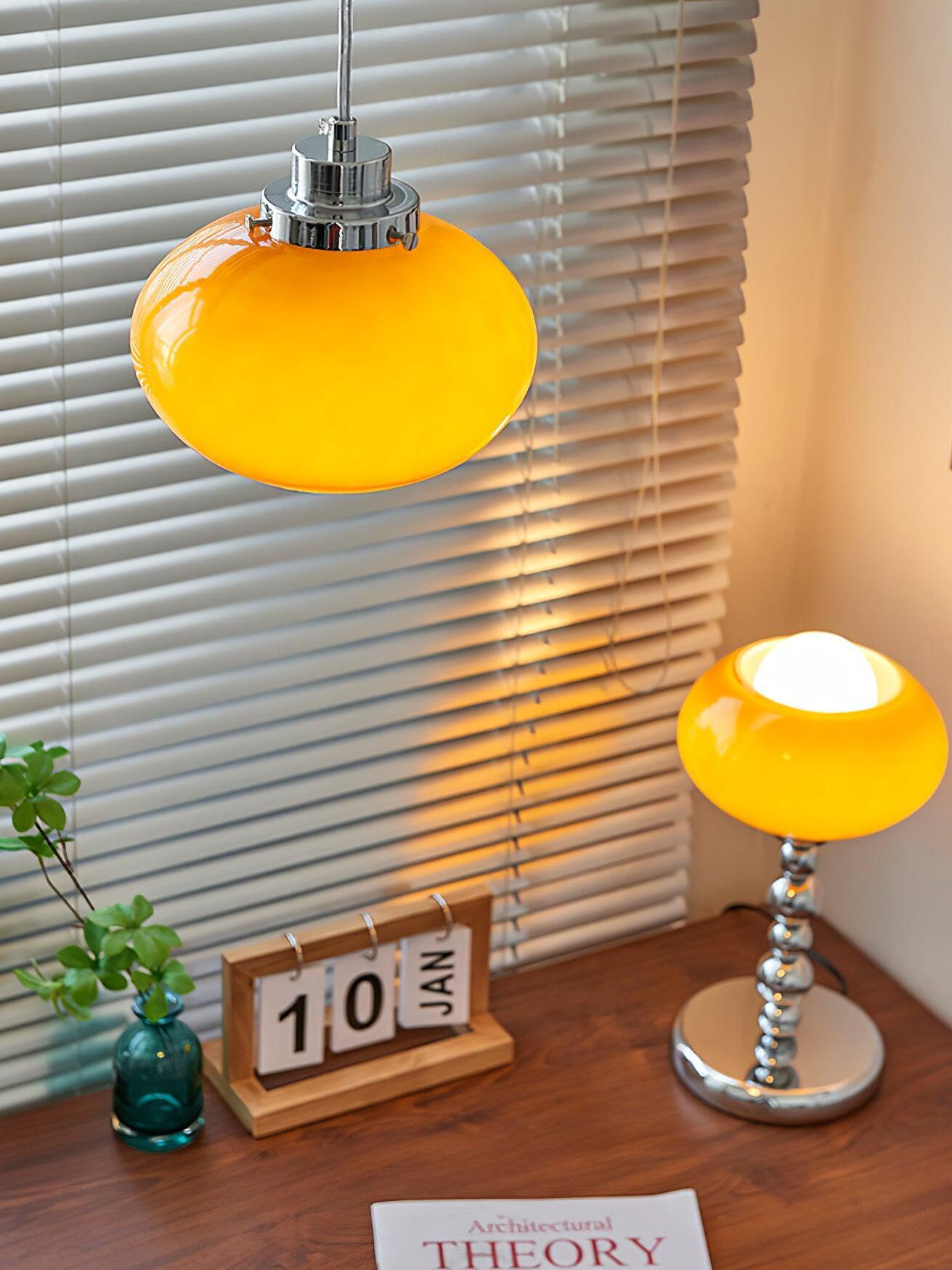 Closer view of the lit orange table lamp and separate hanging light, with chrome fittings beside blinds and a plant