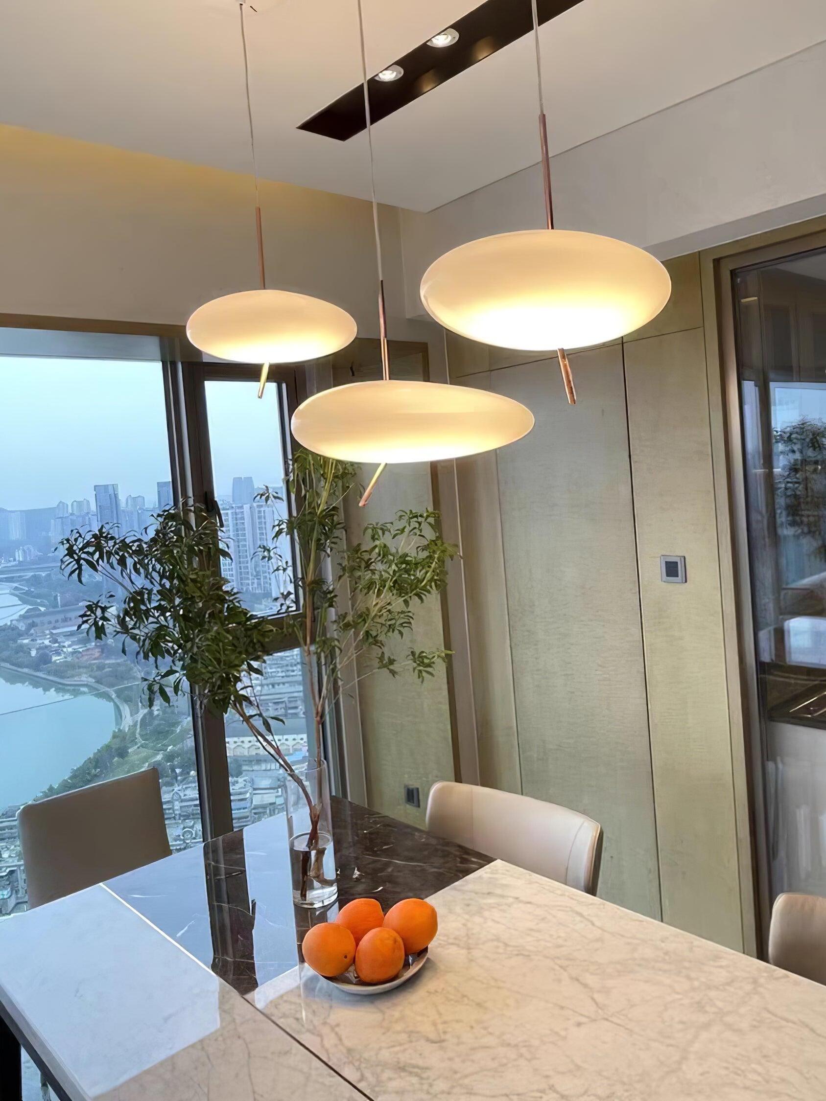 Three glowing white pendant shades at different heights above a table with oranges and a vase