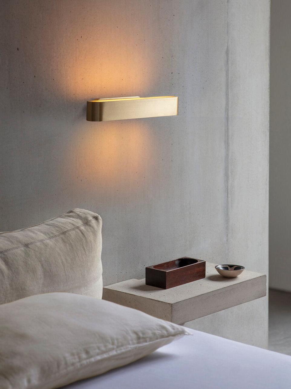 Horizontal gold-tone wall bar casting glow above and below beside a bed with pale bedding and shelf with wood box and bowl