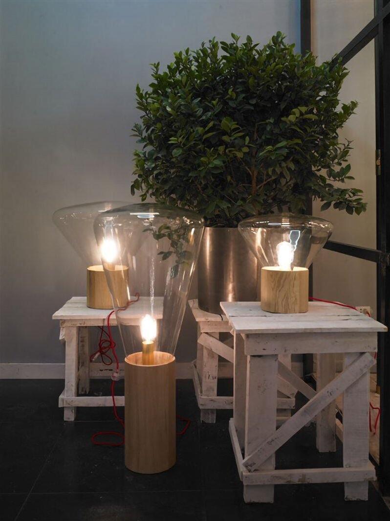 Three clear glass and wood lamps arranged on pale stands and floor near plant
