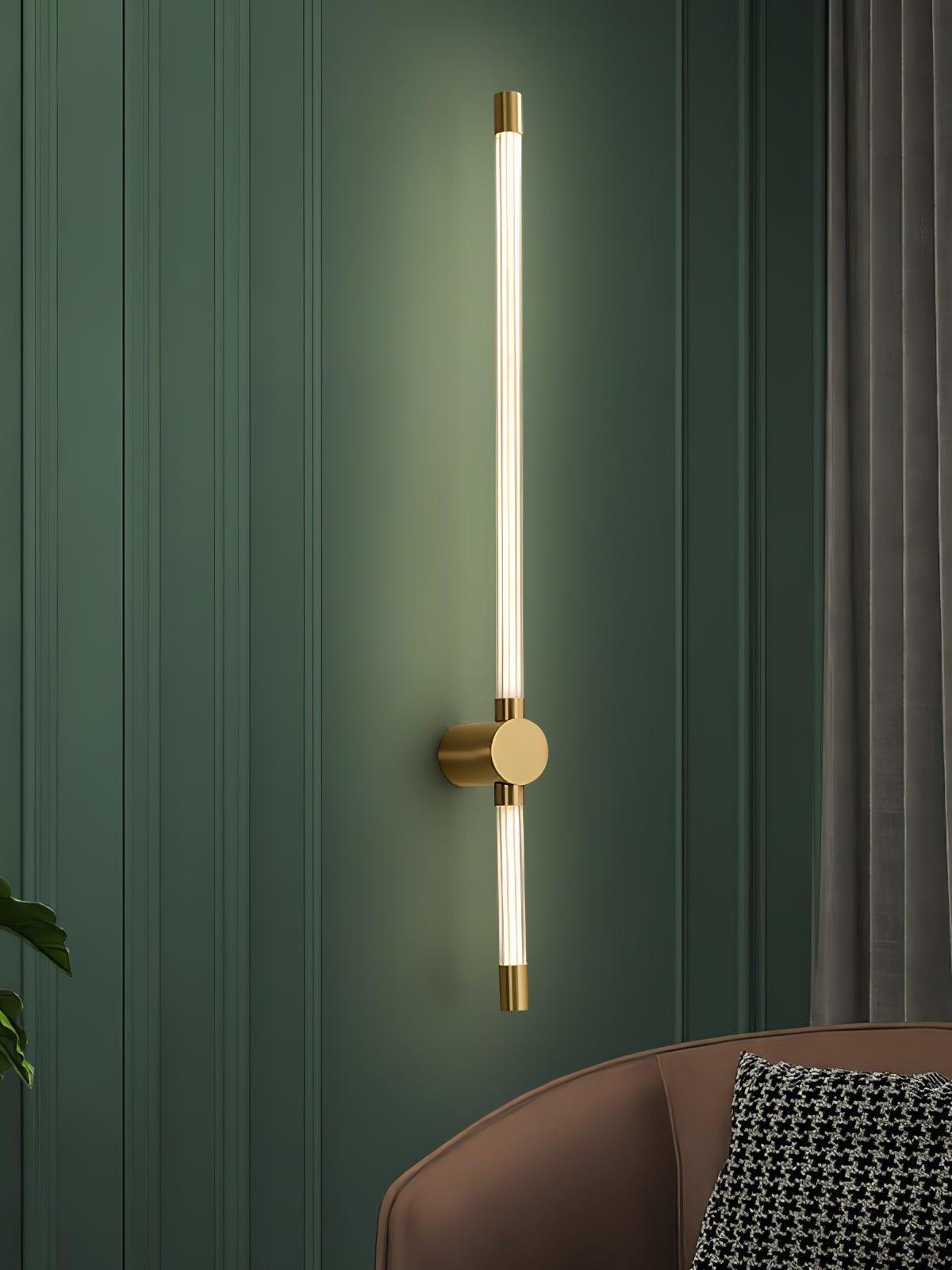 Glowing brass-tone tube wall sconce on a dark green paneled wall beside a brown sofa