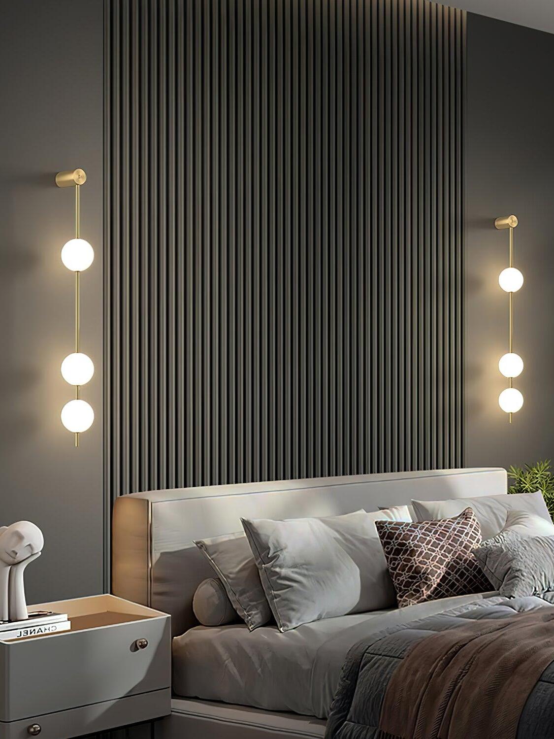 Two lit three-globe wall lamps beside a bed and black fluted panel, with a sculptural bedside decoration
