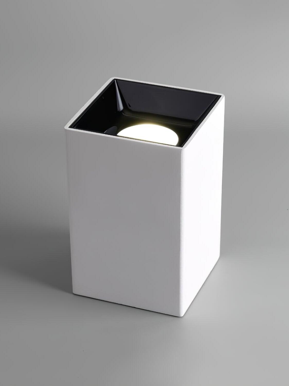 White square spotlight standing with its lit circular aperture facing upward inside a black recessed cup