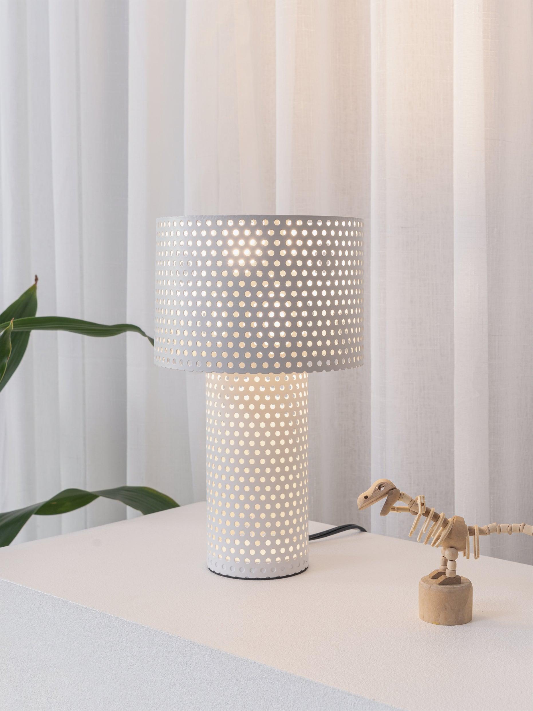 White table lamp with perforated drum beside plant