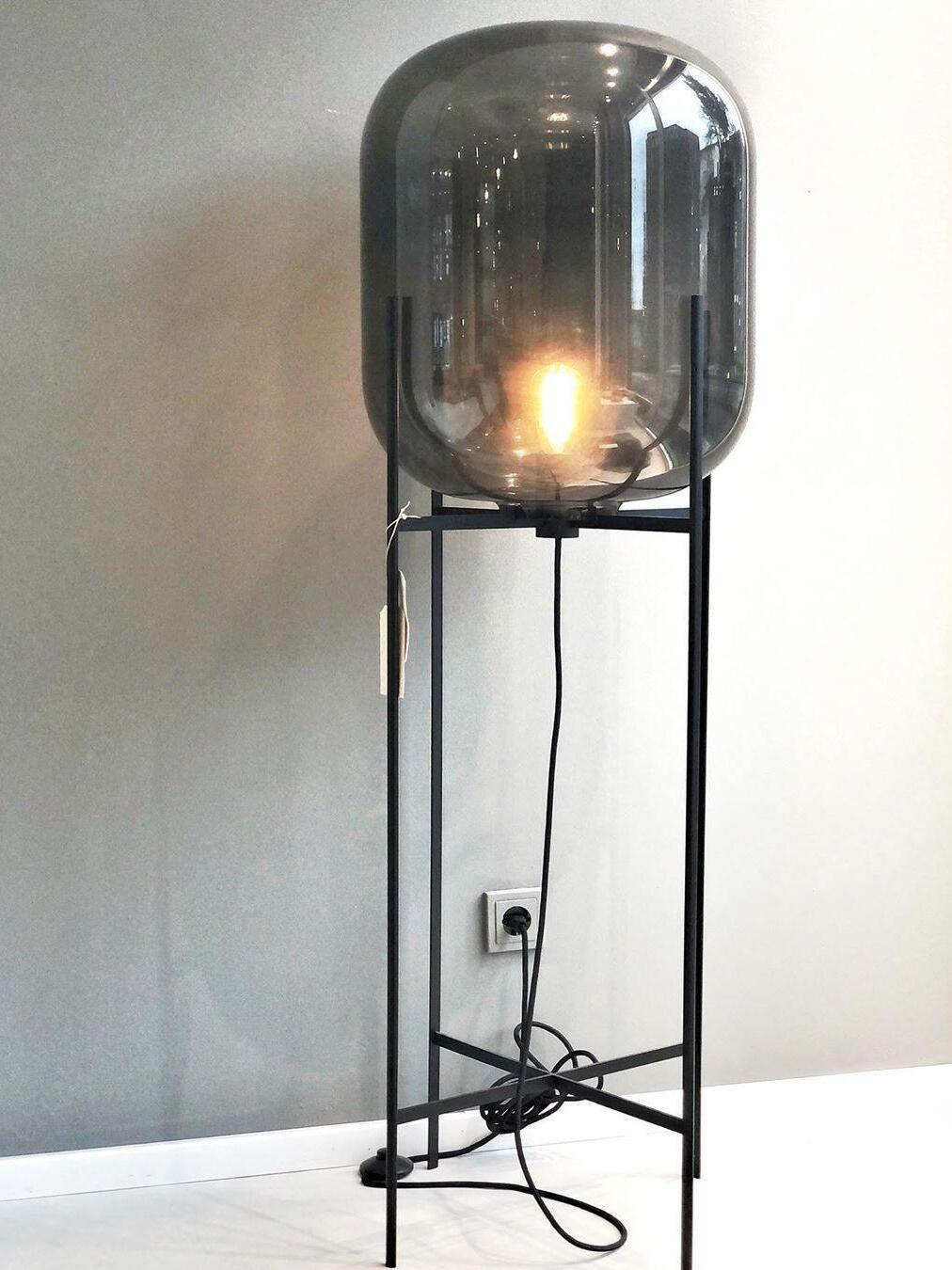 Grey glass Oda floor lamp with a glowing bulb and cord by a wall outlet.